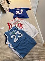 Three basketball jerseys laid out on carpet floor: Hampton 27 in navy blue, Wade 3 in white and red, and Jordan 23 light blue North Carolina jersey.