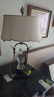 Full view of vintage glass statuette lamp on black marble base with metal frame and beige scalloped fabric shade, showing overall condition and design.