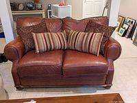 Front view of brown leather loveseat with two paisley and two striped fabric pillows, showing general condition and design.
