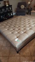 Full view of a large, beige, tufted fabric square Ottoman with wooden legs in a living room setting.