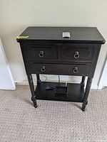 Front view of black wood side table with three drawers showing metal ring pulls and lower shelf with some electronic items placed beneath.