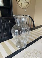 Front angle view of the clear glass Tiffany & Co vase on a table with striped and floral print tablecloth, showing the bulbous fluted body and wide flared rim.