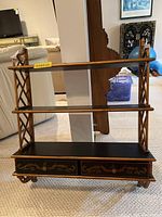 Front view of painted wall shelf showing two shelves, lattice side panels, and two drawers with gold decorative scrollwork and brass knobs.