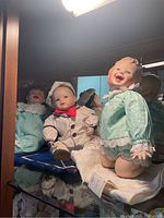 Three Ashton Drake dolls displayed on glass shelf with mirror background. Dolls include a smiling baby doll in turquoise outfit, a baby doll in blue dress, and a boy doll in sailor outfit.