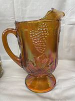 Side view of marigold carnival glass footed pitcher showing grape cluster and leaf pattern with handle.