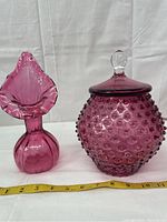 Image showing cranberry glass hobnail covered jar with lid and cranberry glass vase with fluted top side by side against a white background with measuring tape.