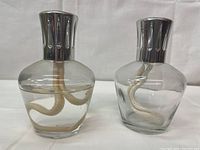 Two clear glass Lampe Berger fragrance oil lamps with silver metal lids and white braided wicks inside. Both lamps have rounded tops and smooth glass bodies.