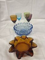 Photo showing amber, blue, green, and purple coloured glass goblets alongside blue scalloped bowls and an amber star-shaped bowl.