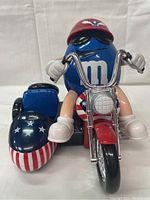 Front view of the blue M&M character riding a motorcycle with American flag themed sidecar, showing helmet and detailed handlebars.