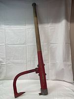 Vintage log roller standing upright, showing wooden handle and red metal body with attached hook.