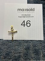 Small gold crucifix pendant placed on a gray textured surface with MaxSold lot number card visible.