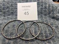 Image showing all three silver bangles together, placed on a gray textured fabric surface with a MaxSold auction tag in the background.