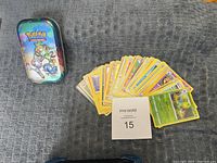 Metal tin with Pokémon design alongside a spread of assorted Pokémon cards on a gray surface, with a MaxSold lot number tag.