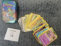 Pokémon metal tin can with assorted Pokémon trading cards fanned out in front, showing a variety of card designs and Pokémon characters.