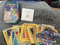 Colorful Pokemon-themed tin can with a stack of assorted Pokemon cards next to it on a gray cloth surface.