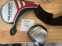 TaylorMade Burner 3 wood club head with 15-degree loft shown with a black, white and red head cover placed on a wooden surface.