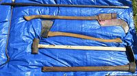 Photo shows five hand tools on a blue tarp: two axes, a sledgehammer, a pry bar, and a pickaxe.