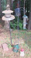 Photo showing multiple bird feeders hanging on black metal shepherd's hooks in an outdoor wooded setting, includes variety of feeder styles and colors including green, black, and white snowman shape.