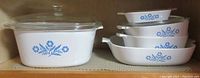 Photo showing 5 CorningWare covered baking dishes with blue daisy design stacked on a shelf, including one large square dish and four smaller dishes with handles/spouts