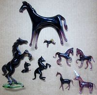 Photo of all nine glass horses arranged on a beige surface showing varying sizes and shapes, many with black or dark glass and some purple accents.
