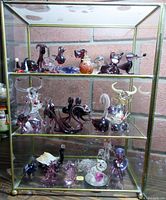 Three-shelf glass cabinet containing multiple glass miniatures of animals, including bulls with horns, swans, dogs, and a perfume bottle on middle shelf.