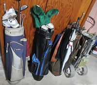 Three golf bags against a wooden door; one contains white golf shoes and clubs, the middle one has green plush head covers and clubs, the third bag has an umbrella and clubs