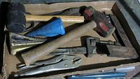 Box of heavy-duty hand tools showing two large hammers, two wedge stamping tools with wooden handles, blue funnel, monkey wrench and vice grips.