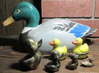 Seven duck figurines including a large ceramic mallard, four brass ducks, and two yellow ceramic ducks side by side.