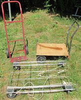 Three carts placed on grassy ground including red two-wheel dolly, wood platform push cart, and wire cart with ropes.