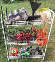 Three-tier white wire shelving rack outdoors holding multiple hardware and tool items including bottles, funnel, jumper cables, and extension cord.