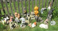 Full view of approximately 20 yard figurines in grass against a fence, including garden gnome, pumpkins, cats, deer, raccoons, squirrel, fox, dogs, and other animals in various materials and colors.