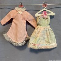 Two Barbie doll dresses hanging on clips. One pink and white pinstriped with lace collar and hem, the other watercolor pastel strapless with pink and white bows and lace detail.