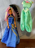 Princess Jasmine doll wearing blue and gold dress alongside green jumpsuit outfit hanging on a hanger.