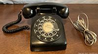 Front angled view of vintage black rotary dial telephone with handset and visible cord on wooden surface.