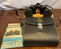 Black Tasco binoculars with lens caps placed on a cardboard box atop a wooden surface, with the black leather case and Tasco manual beside them.