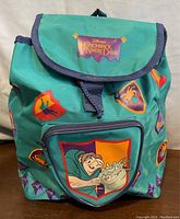 Front view of vintage Disney The Hunchback of Notre Dame backpack predominantly teal with character graphics on front pocket and banners printed all over. Purple trim and Disney logo patch on flap.