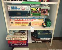 Shelf with various board games stacked vertically and horizontally including Super Stamper, Trivial Pursuit, Admirals, Battleship, Stop Thief, Careers, Tour de Force, Le Bourreau, Othello, Trac 4, Double Line, Junior Trivia, and a spherical puzzle ball.