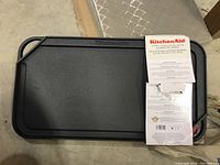 Cast iron grill/griddle pan with packaging and instructional papers on top, showing flat cooking surface and handles.