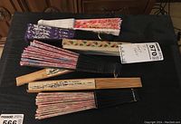 Seven assorted hand fans displayed partially open and closed on a dark surface.