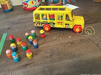 Yellow Fisher Price pull along school bus with 12 small wooden peg doll figures beside it on a wooden floor.