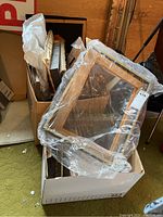 Cardboard box filled with various picture frames and wrapped framed art pieces, showing wooden frames and glass protection covers.