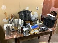 Photo showing black speckled enamelled pickling pots, metal pasta maker, springform pan, metal shaker, metal grater, kitchen utensils and other kitchen items on a table.