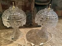 Pair of clear glass lamps with dome-shaped shades featuring grid patterns and floral motifs on the shades and bases, brass finials on top, white cords with switches, sitting on carpet.