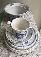 Set of stacked dinner and side plates, creamer jug on top, bowl, and serving dish in background showing blue floral motives on white ceramic.