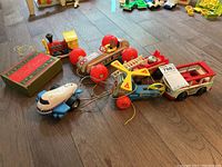 Photo showing a collection of vintage wooden pull toys including a Tonka plane, race car, fire engine, mini bus, train engine, and box of wooden buildings on a wood floor.