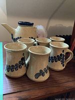 Photo showing six ceramic mugs and one teapot all cream-colored with dark blue leaf patterns and speckled finish