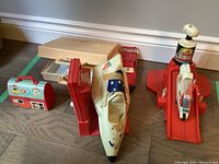 Grouping showing empty Fisher Price medical kit case, barn lunch box and thermos, space shuttle, and red launcher with Six Million Dollar Man figure in vehicle.