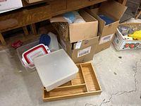 Wide view showing assorted kitchenware including boxes with containers, collapsible colander, and bamboo drawer organizer on concrete floor under a workbench