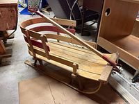 Side view of vintage wooden sled showing wood slat seating, side rails, metal skis, and wooden steering handle with red paint.