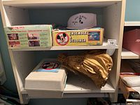 Two shelf section showing several items including the boxed Mickey Mouse Club drawing desk, a gold angel figurine, a gray hat, a closed styrofoam container, and a Radio Shack truck box on top shelf.